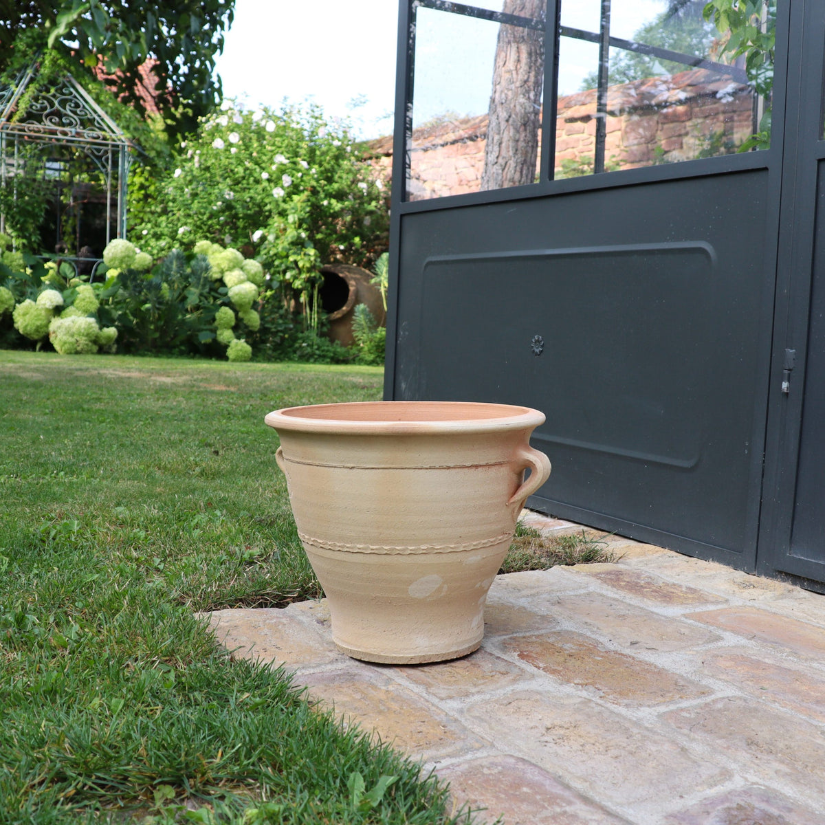 Im Garten steht ein Terrakotta-Blumentopf „Rosa“ (H=25 cm) auf einem Steinweg neben einer schwarzen Metall- und Glaskonstruktion. Der üppige Garten bietet grünes Gras, Büsche mit weißen Blüten und im Hintergrund einen dekorativen Metallpavillon. Der Topf ergänzt die ruhige Umgebung wunderbar.