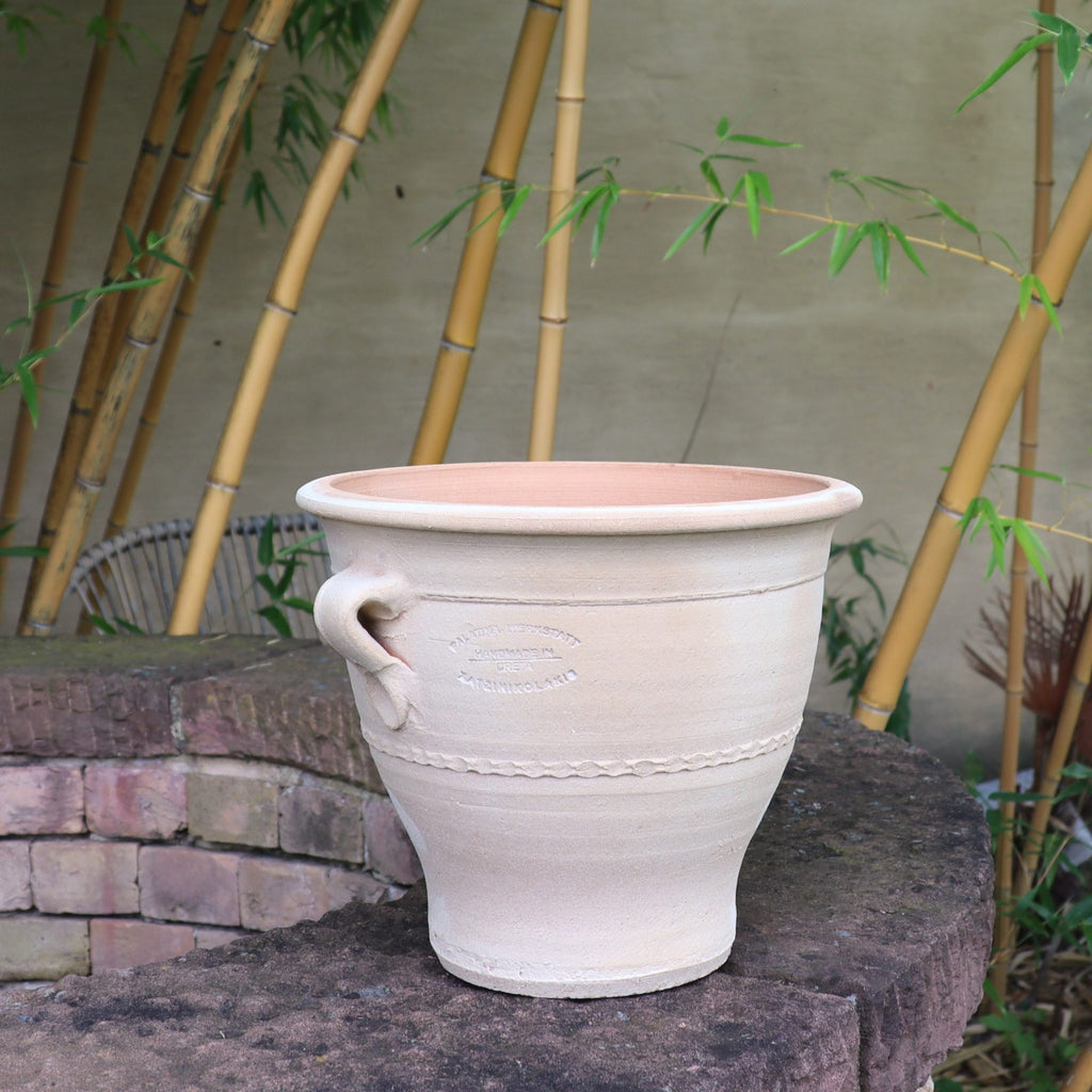 Ein großer, cremefarbener Keramiktopf mit dekorativer Struktur und seitlichem Griff steht auf einem Ziegelsims. Daneben sorgt der 25 cm hohe Blumentopf „Rosa“ aus Terrakotta für eine rustikale Note. Im Hintergrund sorgen hohe Bambusstäbe und helle Wände für eine ruhige Gartenatmosphäre.