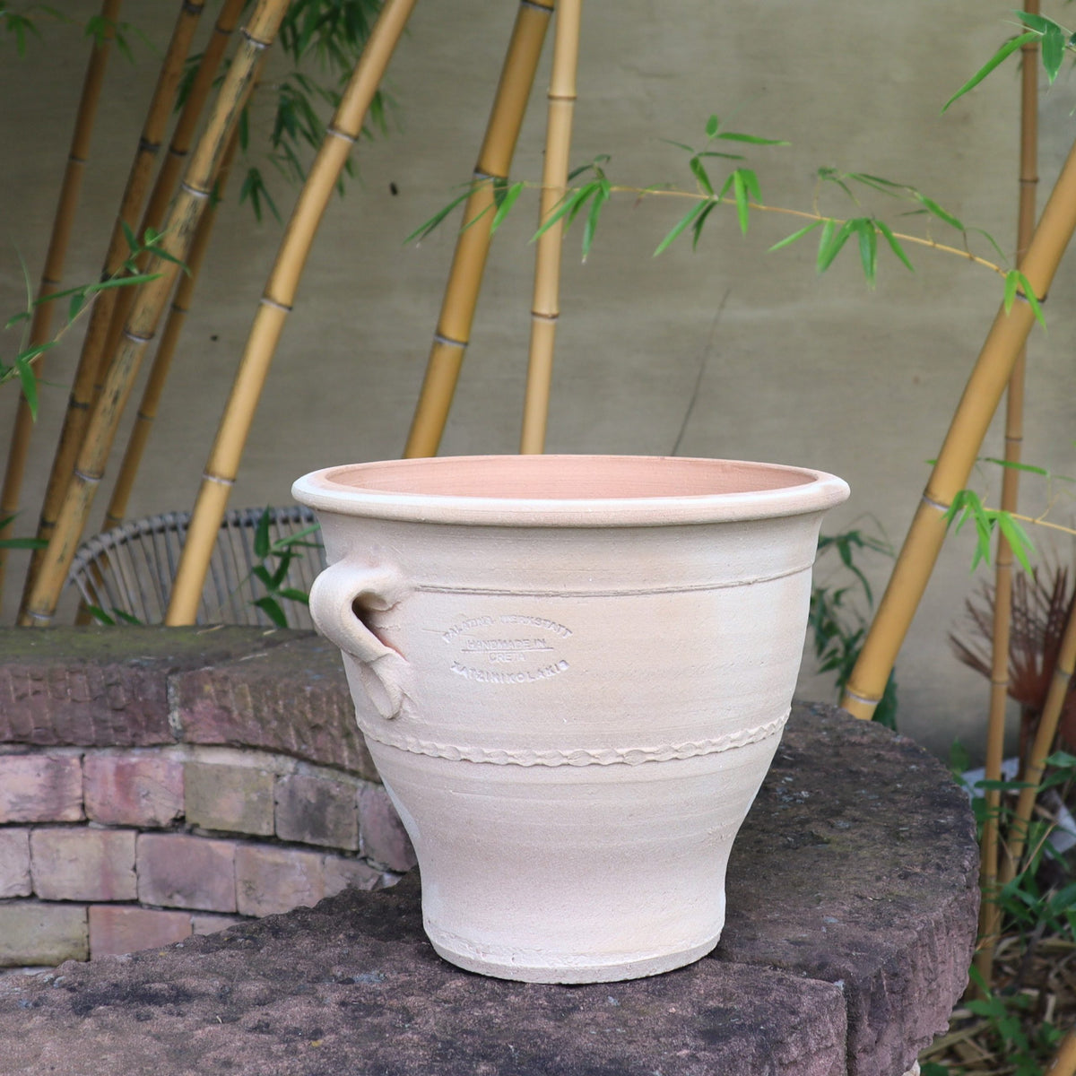 Ein großer, cremefarbener Keramiktopf mit dekorativer Struktur und seitlichem Griff steht auf einem Ziegelsims. Daneben sorgt der 25 cm hohe Blumentopf „Rosa“ aus Terrakotta für eine rustikale Note. Im Hintergrund sorgen hohe Bambusstäbe und helle Wände für eine ruhige Gartenatmosphäre.