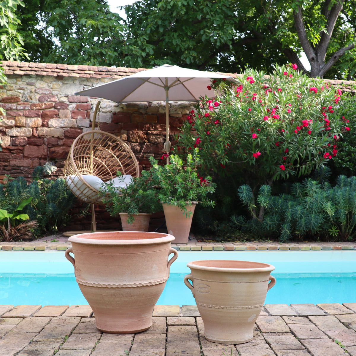 Eine ruhige Szene am Pool zeigt zwei große Terrakotta-Blumentöpfe „Rosa“, H=25 cm, im Vordergrund auf einer Steinterrasse. Im Hintergrund tragen ein Korbstuhl, ein Sonnenschirm, üppiges grünes Laub und blühende rote Blumen zur ruhigen Gartenatmosphäre bei.