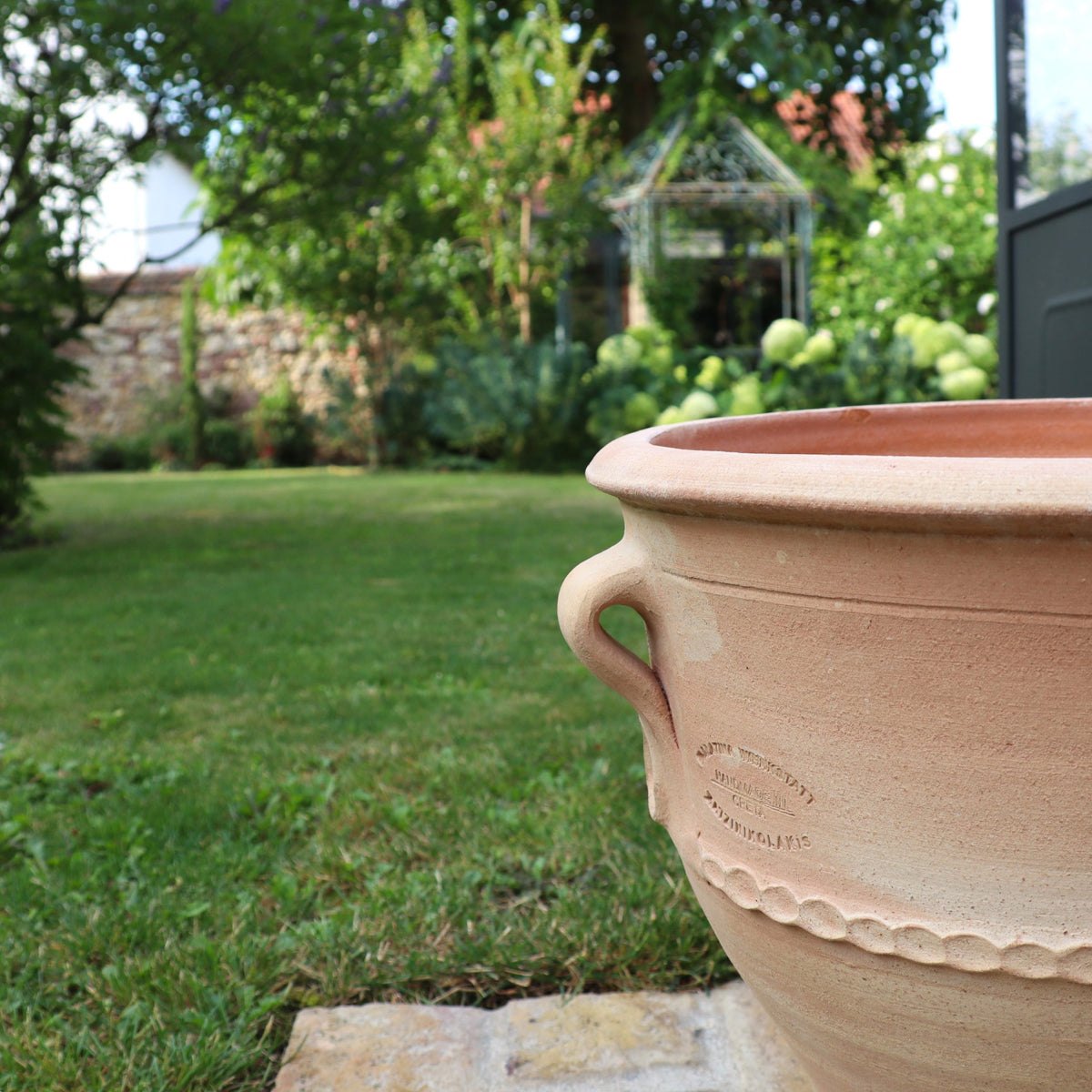 Im Vordergrund steht ein großer Terrakotta-Pflanzkübel „Rosa 2“ (H=35cm) auf einer Steinplatte, während im Hintergrund ein üppiger Garten mit grünen Sträuchern, blühenden Blumen und einem Steinweg zu sehen ist, der zu einem Pavillon mit Metallrahmen führt. Die Szene wird durch natürliches Tageslicht beleuchtet.
