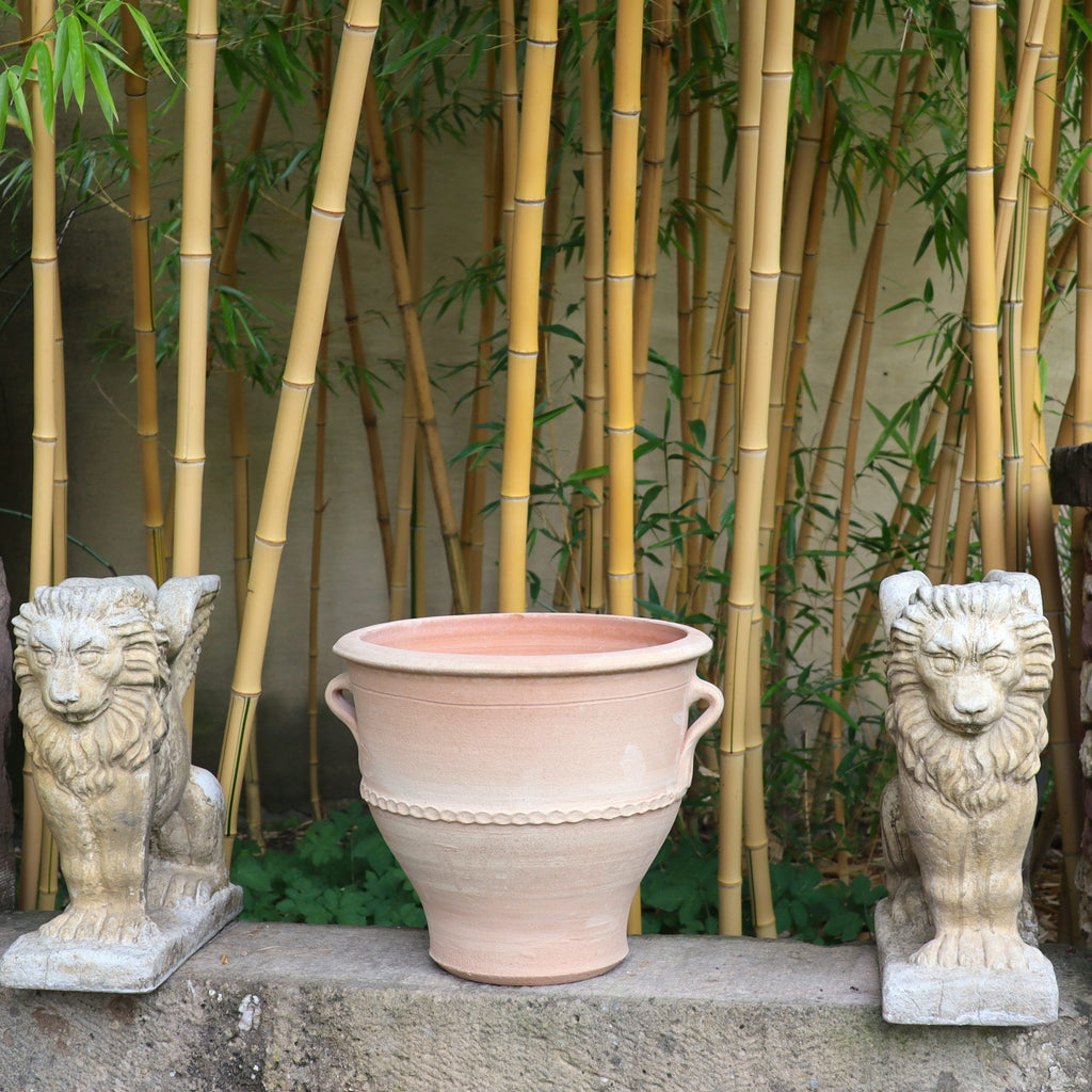 Ein Terrakotta-Pflanzkübel „Rosa 2“ (H=35 cm) steht zwischen zwei Löwenstatuen aus Stein auf einem Steinsims. Der Hintergrund besteht aus hohen Bambusstäben, die eine ruhige und natürliche Atmosphäre schaffen.