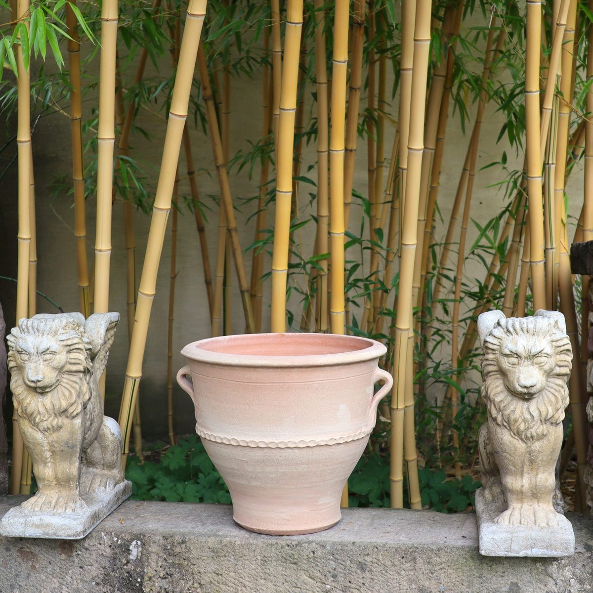 Ein Terrakotta-Pflanzkübel „Rosa 2“ (H=35 cm) steht zwischen zwei Löwenstatuen aus Stein auf einem Steinsims. Der Hintergrund besteht aus hohen Bambusstäben, die eine ruhige und natürliche Atmosphäre schaffen.