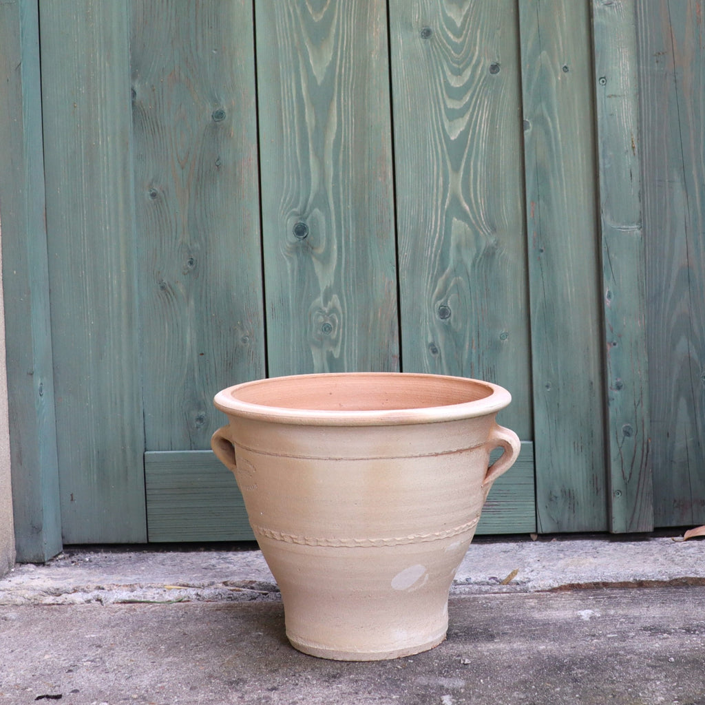 Vor einer grünen, verwitterten Holztür steht der Terrakotta-Blumentopf „Rosa“ (H=25 cm) mit Griffen auf beiden Seiten auf einer Betonfläche. Seine große, beige Terrakottaform und sein rustikales Design bilden einen auffälligen Kontrast zum strukturierten, gealterten Holzhintergrund und machen ihn zu einer perfekten Ergänzung für Rosenblüten.