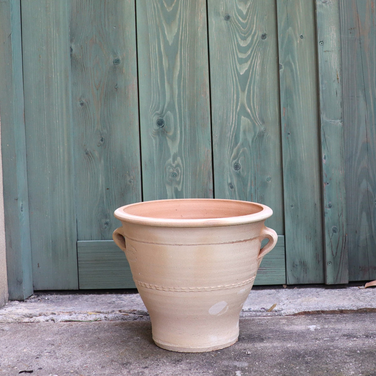 Vor einer grünen, verwitterten Holztür steht der Terrakotta-Blumentopf „Rosa“ (H=25 cm) mit Griffen auf beiden Seiten auf einer Betonfläche. Seine große, beige Terrakottaform und sein rustikales Design bilden einen auffälligen Kontrast zum strukturierten, gealterten Holzhintergrund und machen ihn zu einer perfekten Ergänzung für Rosenblüten.