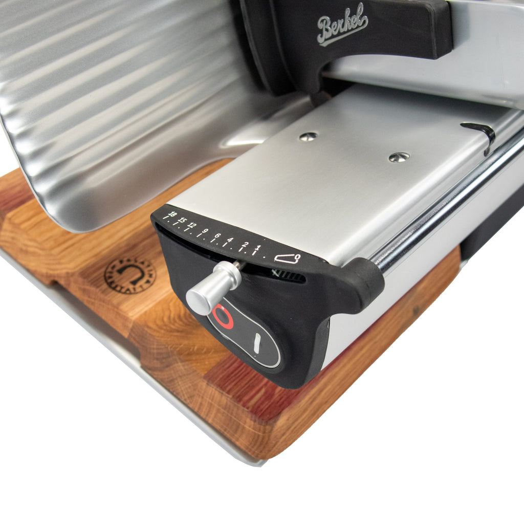 Nahaufnahme der Berkel Home Line 200 Plus schwarz matt Aufschnittmaschine mit handgefertigtem Schneidebrett & Wellenschliff-Bausatz für Brot auf Holz, mit sichtbarem Einstellknopf, nummeriertem Drehregler und Berkel-Logo.