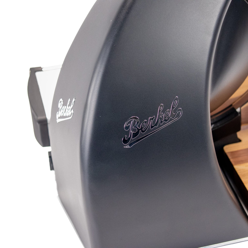 Nahaufnahme der Berkel Home Line 250 Plus Schwarz MATT Aufschnittmaschine - Neues Modell, die das geschwungene Gehäuse mit silbernem "Berkel"-Logo und einen Teil des einzigartigen, handgefertigten Schneidebretts auf der rechten Seite zeigt.