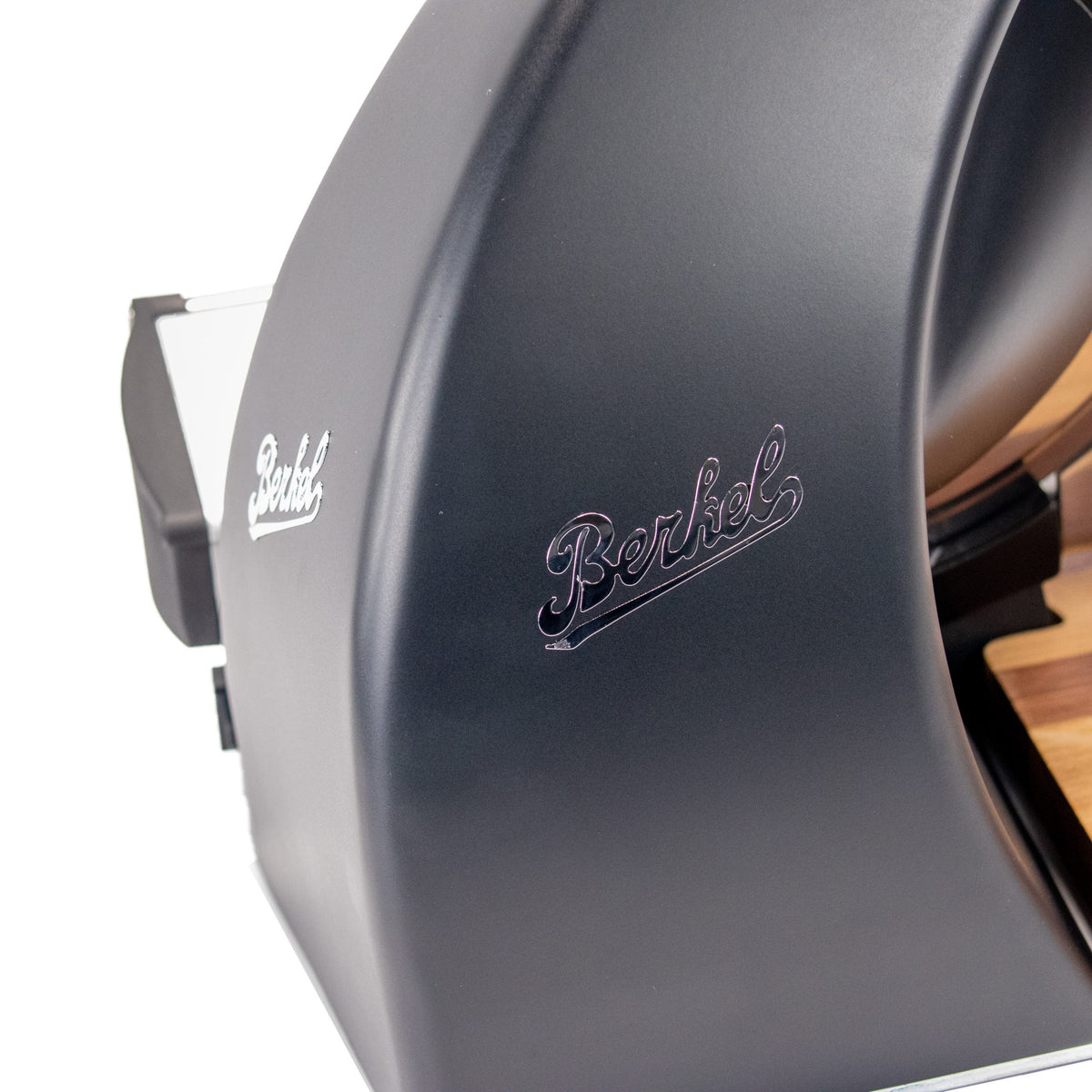 Nahaufnahme der Berkel Home Line 250 Plus Schwarz MATT Aufschnittmaschine - Neues Modell, die das geschwungene Gehäuse mit silbernem "Berkel"-Logo und einen Teil des einzigartigen, handgefertigten Schneidebretts auf der rechten Seite zeigt.