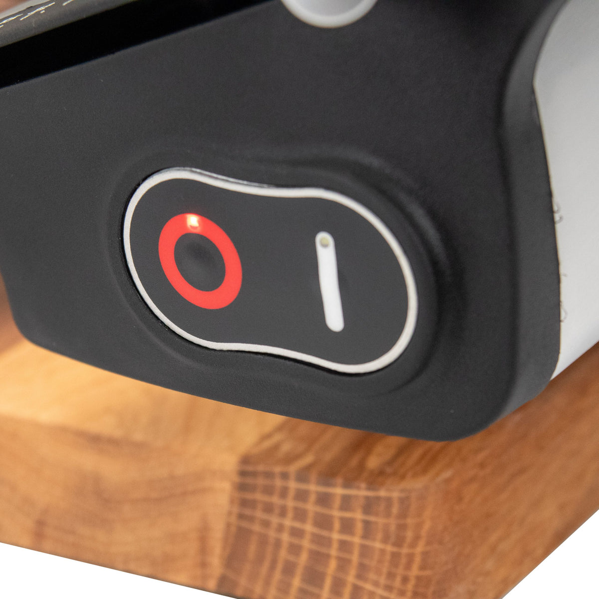 Eine Nahaufnahme der Berkel Home Line 250 Plus Schwarz MATT Aufschnittmaschine - Neues Modell - zeigt den schwarzen Ein/Aus-Schalter mit roter Kontrollleuchte, der an der Maschine angebracht ist, die auf ihrem einzigartigen 100% handgefertigten Schneidebrett ruht.