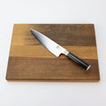 Ein Kai Shun Classic DM-0706 Kochmesser mit schwarzem Griff liegt horizontal auf einem übergroßen Holzschneidebrett aus Fassholz mit den Maßen 40x30 cm. Das Schneidebrett ist zweifarbig gestaltet mit dunkleren und helleren Holzpartien. Die polierte 20 cm lange Klinge des Messers weist ein dezentes Wellenmuster auf, das sich von einem schlichten weißen Hintergrund abhebt.