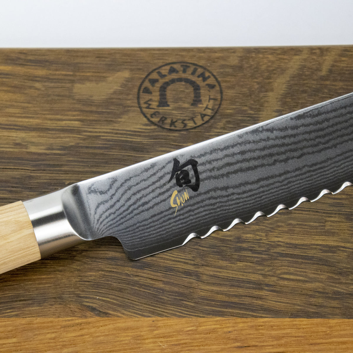 Eine Nahaufnahme eines gezackten Kai Shun Classic White DM-0705W Brotmessers mit einem gewellten Damaszenermuster auf seiner 23,0 cm langen Klinge, das auf einem 40 x 30 cm großen handgefertigten Schneidebrett ruht. Auf dem Schneidebrett ist ein Logo mit dem Text „Palatina Werkstatt“ zu sehen. Das Messer hat einen hellen Holzgriff.