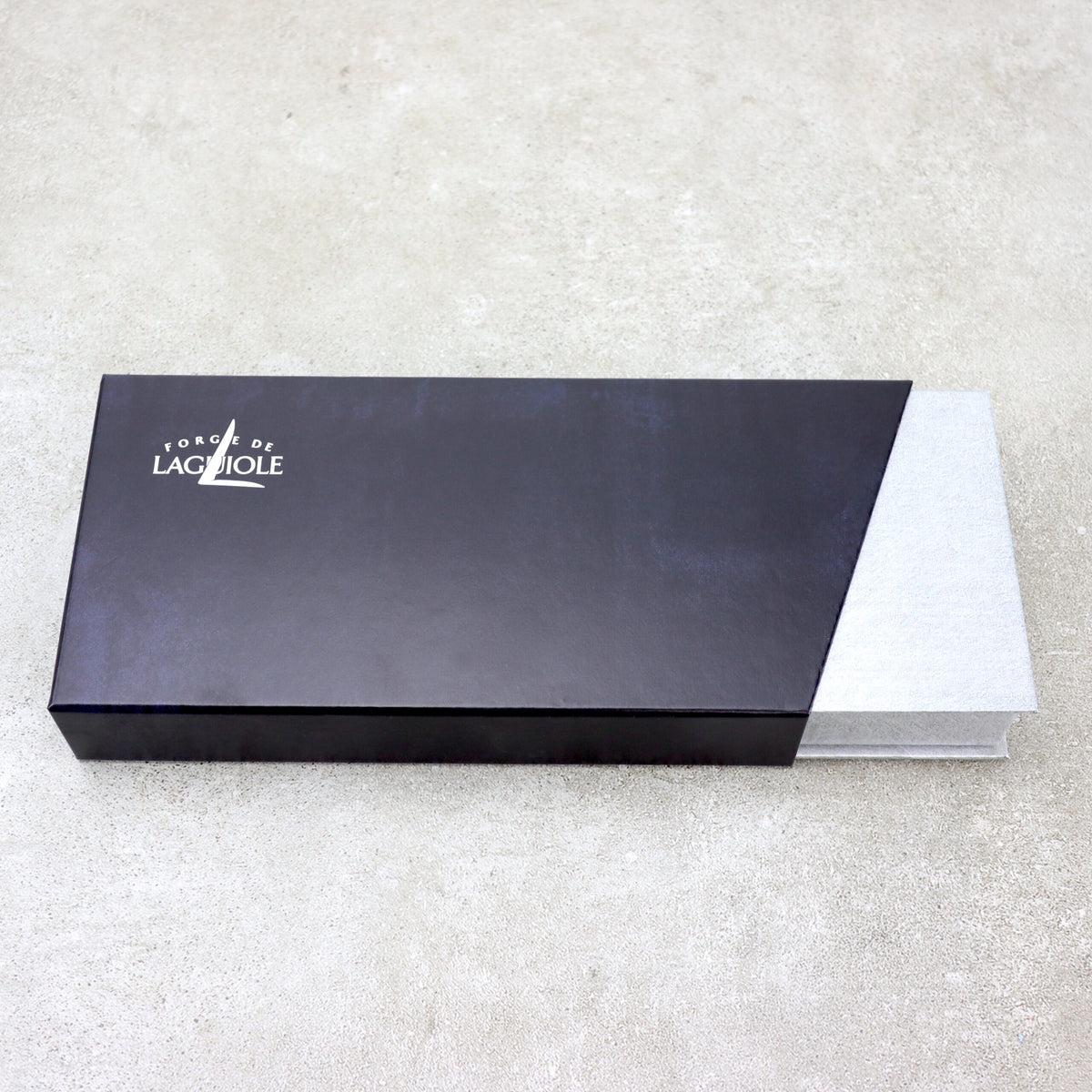 Eine schwarz-graue Box mit dem Text „L'ORGE DE LAGUIOLE“ und einem kleinen Baumlogo in der oberen linken Ecke ruht auf einer hellen Oberfläche. Teilweise geöffnet zeigt sie ihr elegantes silberfarbenes Inneres und lässt die Verfeinerung des Forge De Laguiole Kellnermesser Sommelier | Amourette glänzend im Inneren erahnen.