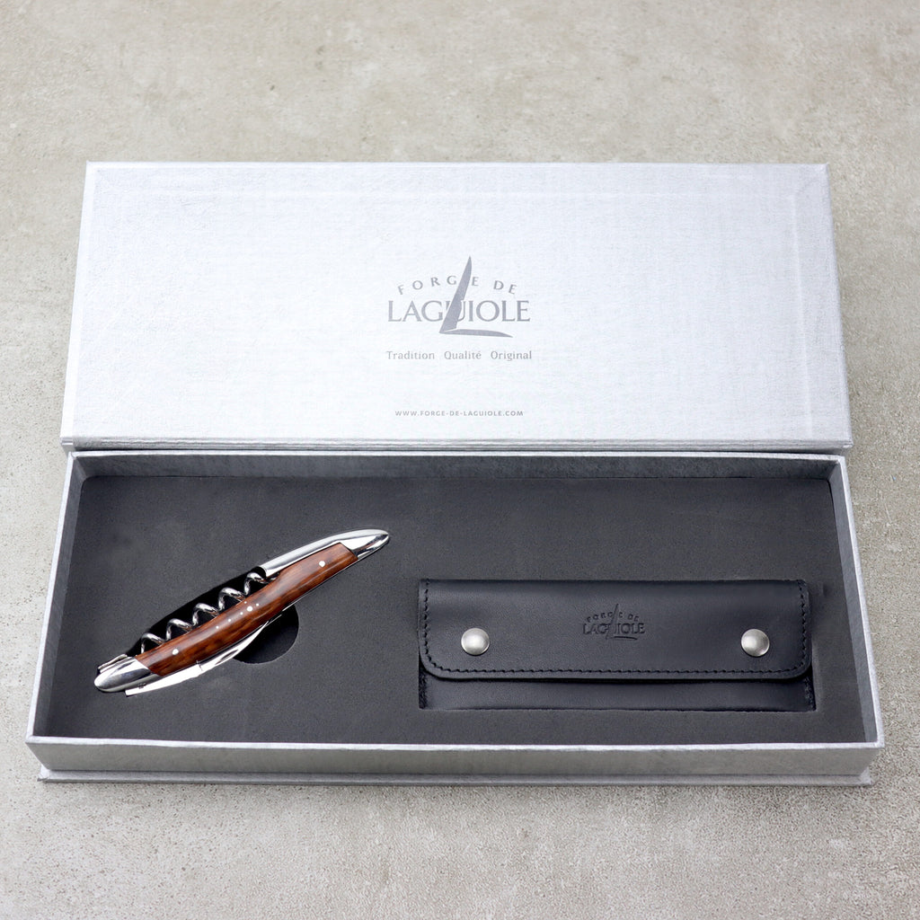 Ein Stift mit schwarzem Griff und silbernen Akzenten wird in einer offenen grauen Schachtel präsentiert. Der Deckel der Schachtel mit der Aufschrift „Forge De Laguiole“ weist auf die Handwerkskunst der Marke hin, ähnlich wie ihr berühmtes Sommeliermesser. Neben dem Stift befindet sich eine passende schwarze Stifthülle aus Leder mit zwei silbernen Knöpfen.