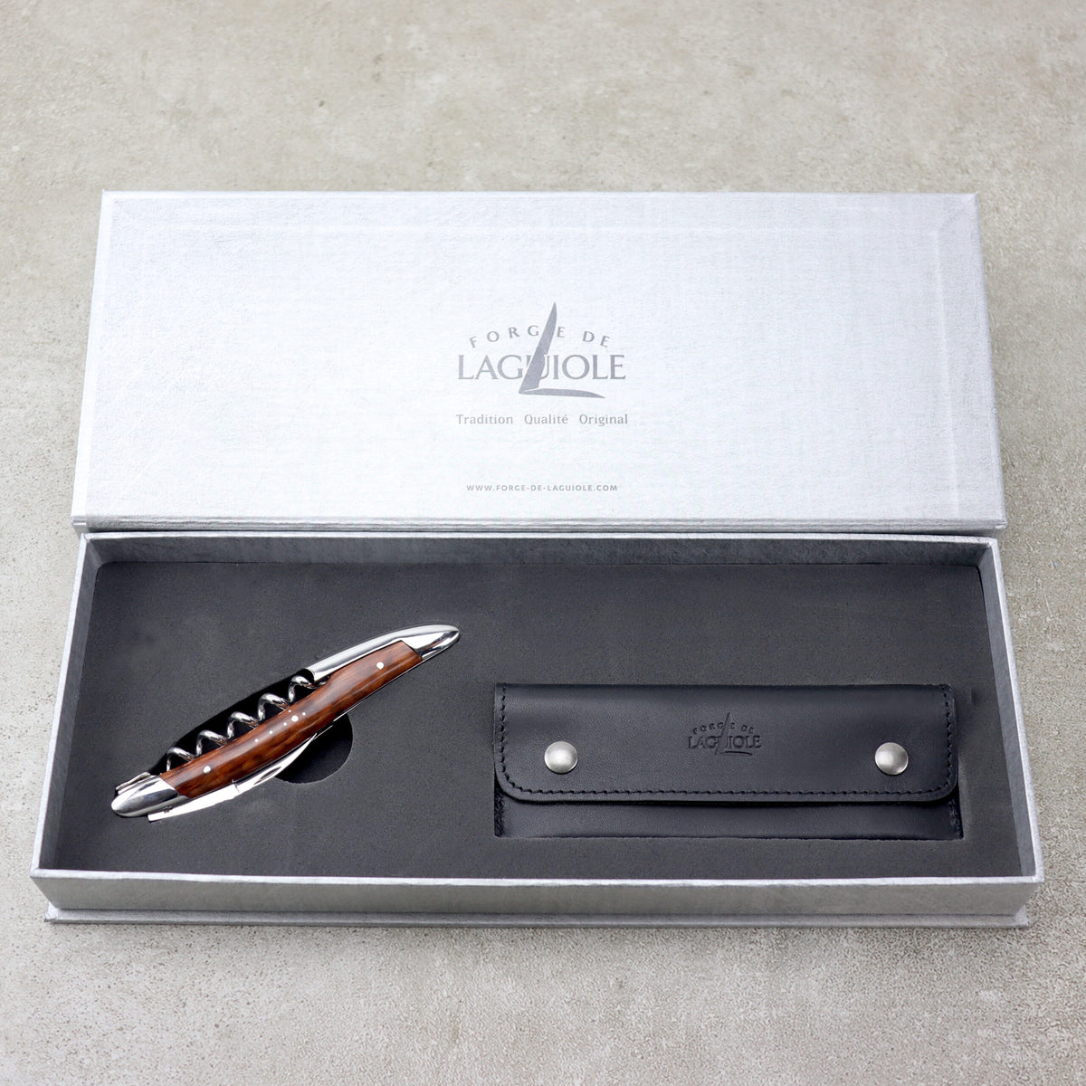 Ein Stift mit schwarzem Griff und silbernen Akzenten wird in einer offenen grauen Schachtel präsentiert. Der Deckel der Schachtel mit der Aufschrift „Forge De Laguiole“ weist auf die Handwerkskunst der Marke hin, ähnlich wie ihr berühmtes Sommeliermesser. Neben dem Stift befindet sich eine passende schwarze Stifthülle aus Leder mit zwei silbernen Knöpfen.