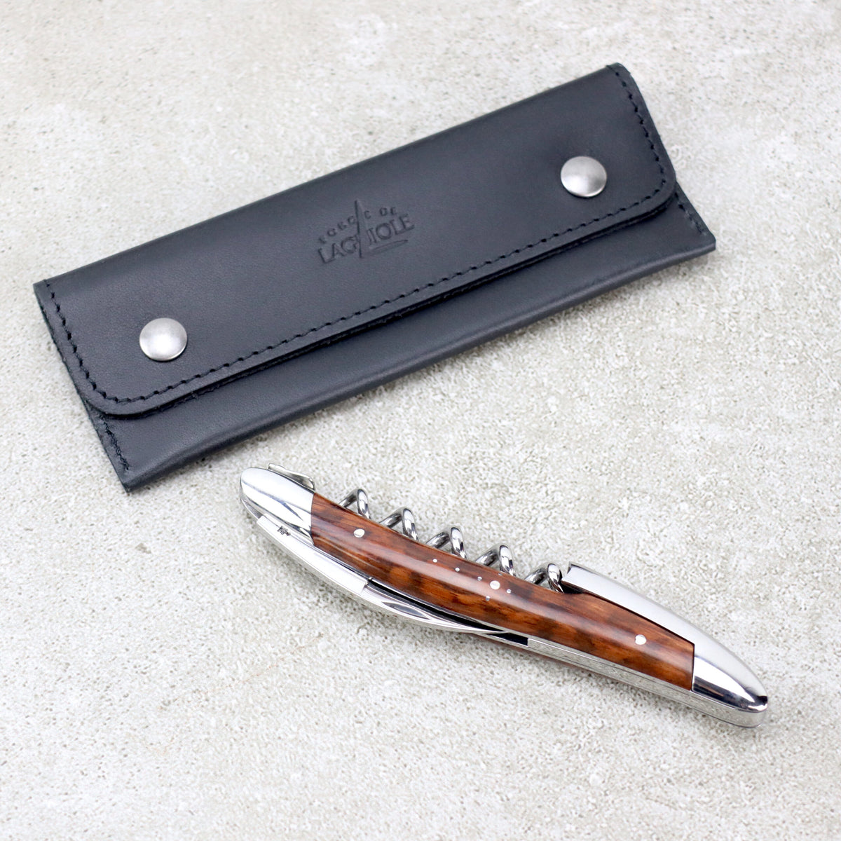 Ein eleganter, faltbarer Weinöffner von Forge De Laguiole Kellnermesser Sommelier | Amourette glänzend mit poliertem Holzgriff liegt neben einer schwarzen Ledertragetasche auf einer strukturierten grauen Oberfläche. Die Tasche hat silberne Knöpfe und eine geprägte Aufschrift von Forge De Laguiole.