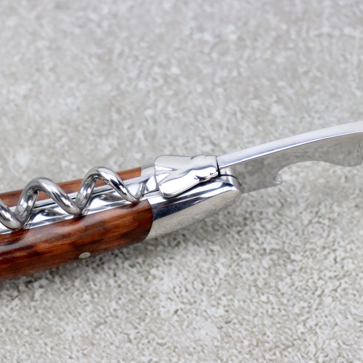 Eine Nahaufnahme des Forge De Laguiole Kellnermessers Sommelier | Amourette glänzend, das seinen Holzgriff und die Edelstahlkomponenten zeigt. Der Korkenzieher liegt auf einer strukturierten grauen Oberfläche, wodurch die spiralförmige Metallspitze und die Flaschenöffnerfunktion hervorgehoben werden – perfekt für jeden anspruchsvollen Sommelier.