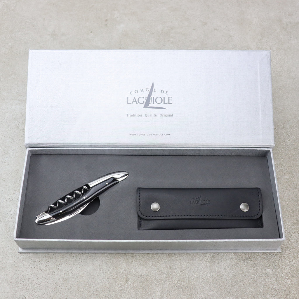 Ein hochwertiges Set aus Kellnermesser Sommelier | Hornspitze glänzend von Forge De Laguiole und Lederetui wird in einer eleganten, offenen grauen Box präsentiert. Der Korkenzieher hat eine polierte Metalloberfläche und einen schwarzen Griff, während das schwarze Lederetui über zwei Druckknöpfe für einen sicheren Verschluss verfügt. Der Innendeckel trägt die Aufschrift „Forge de Laguiole“.