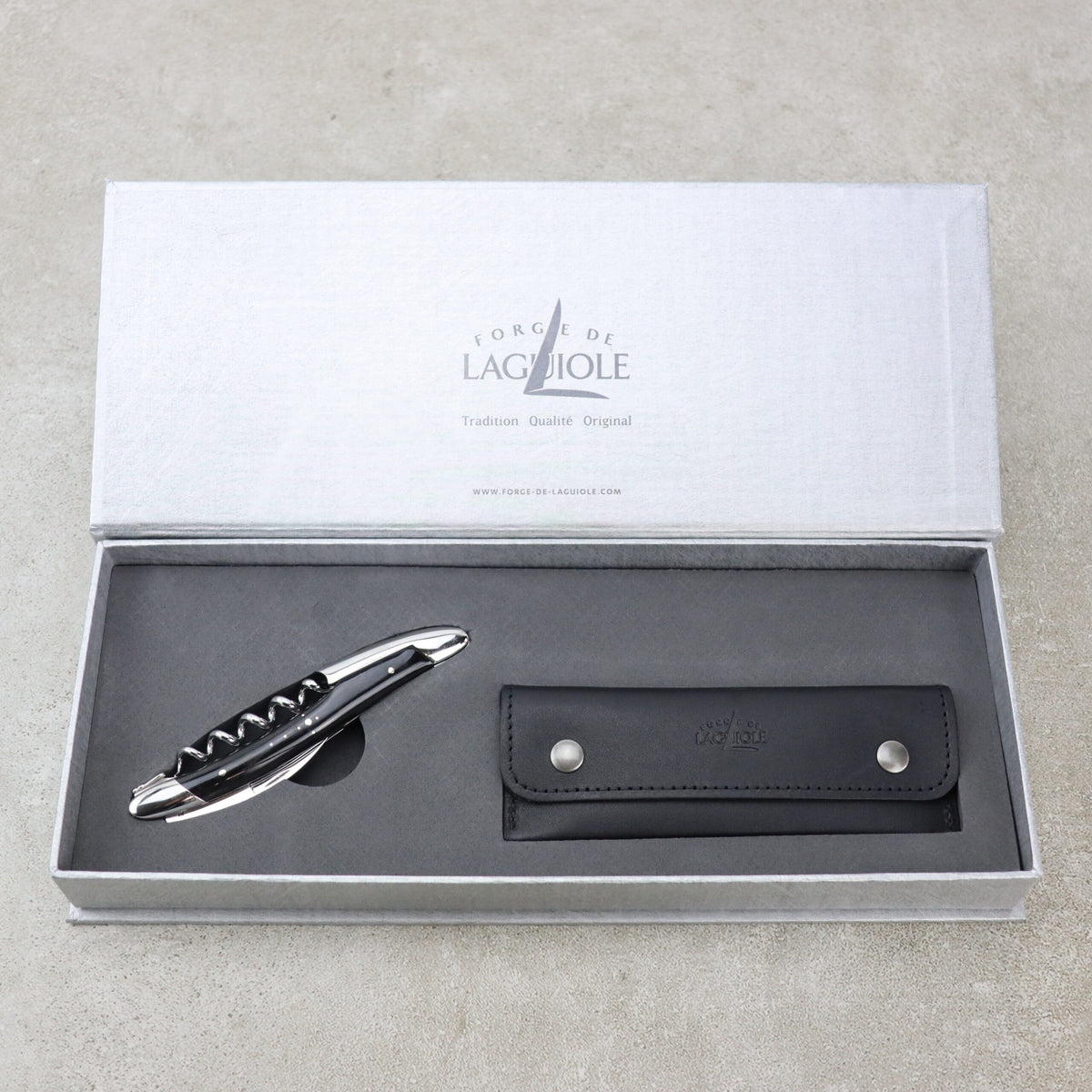 Ein hochwertiges Set aus Kellnermesser Sommelier | Hornspitze glänzend von Forge De Laguiole und Lederetui wird in einer eleganten, offenen grauen Box präsentiert. Der Korkenzieher hat eine polierte Metalloberfläche und einen schwarzen Griff, während das schwarze Lederetui über zwei Druckknöpfe für einen sicheren Verschluss verfügt. Der Innendeckel trägt die Aufschrift „Forge de Laguiole“.