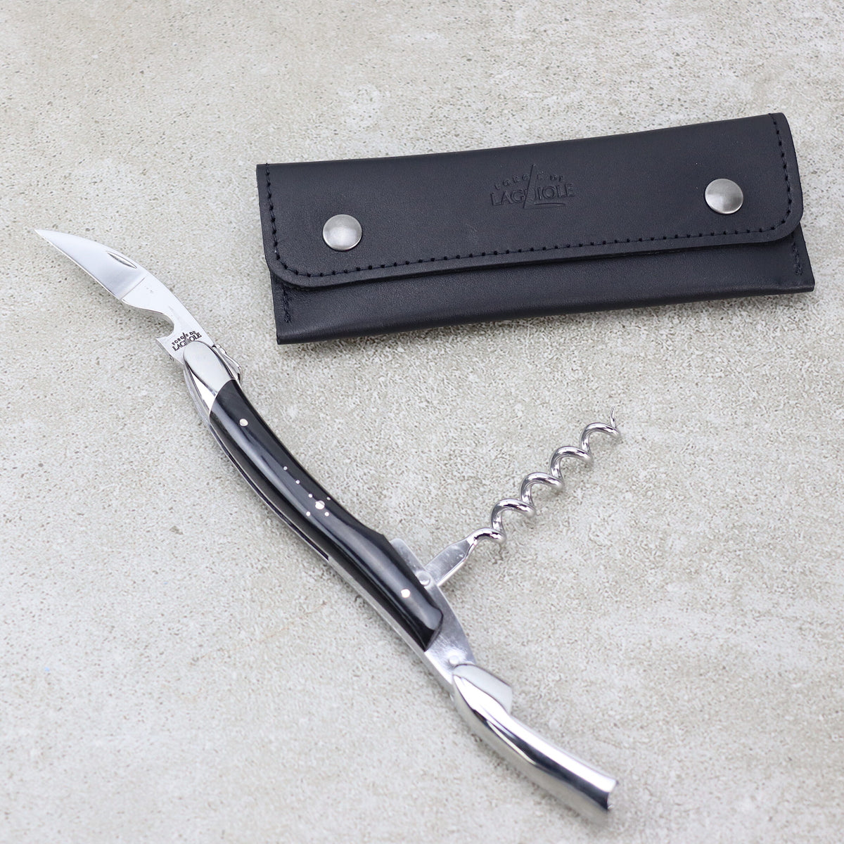 Ein Kellnermesser Sommelier von Forge De Laguiole mit einem eleganten schwarzen Griff und silbernen Akzenten liegt geöffnet auf einer Betonoberfläche. Daneben befindet sich ein schwarzes Lederetui mit Knopfverschluss. Das Messer, das an ein edles Modell mit glänzender Hornspitze erinnert, verfügt über mehrere Werkzeuge, darunter einen Korkenzieher für den anspruchsvollen Sommelier.