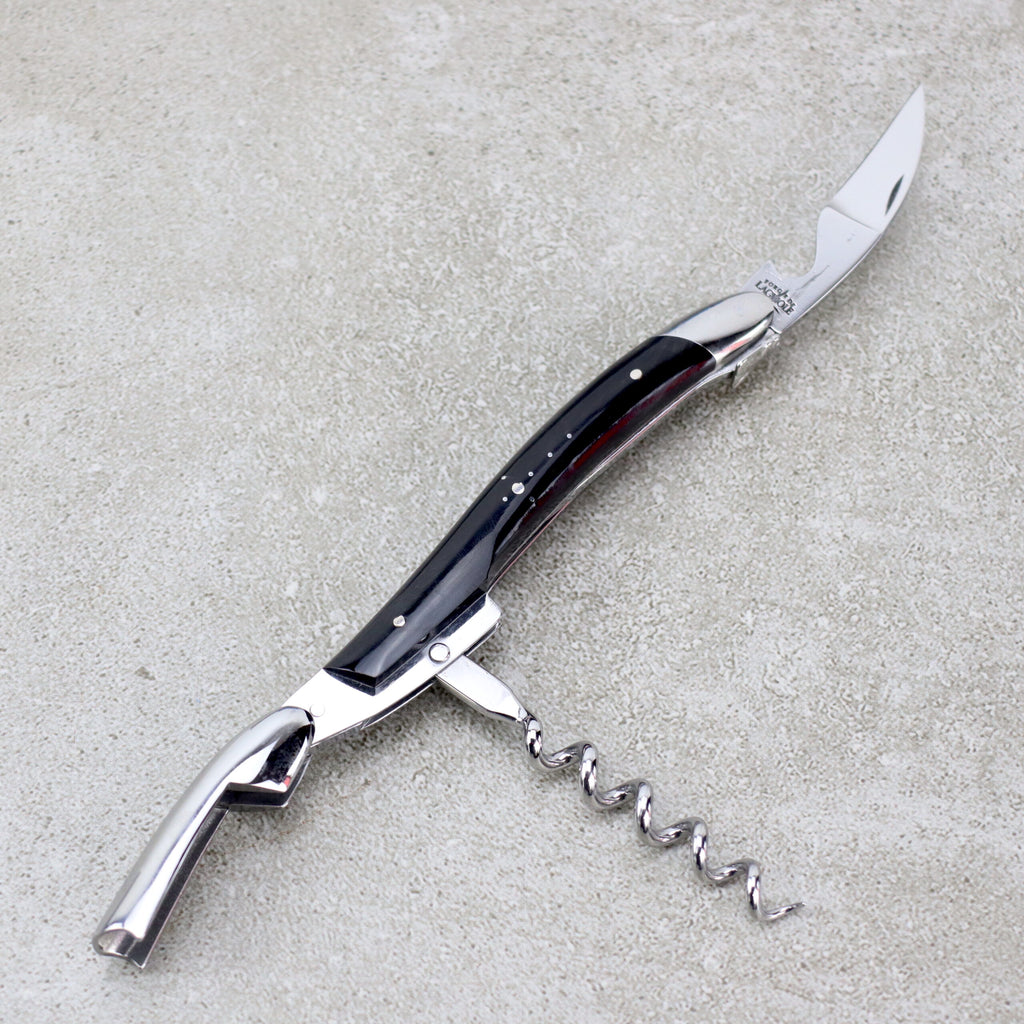 Das Forge De Laguiole Kellnermesser Sommelier | Hornspitze glänzend ist ein multifunktionales Werkzeug mit einer gebogenen Messerklinge, einem ausklappbaren Korkenzieher und einem Flaschenöffner. Der schlanke Griff weist dunkle und metallische Akzente auf und verkörpert die Handwerkskunst von Forge De Laguiole. Dieses exquisite Sommelier-Werkzeug ruht auf einer grauen, strukturierten Oberfläche und ist damit eine unverzichtbare Ergänzung für die Sammlung eines jeden Sommeliers.