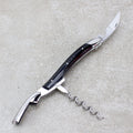 Das Forge De Laguiole Kellnermesser Sommelier | Hornspitze glänzend ist ein multifunktionales Werkzeug mit einer gebogenen Messerklinge, einem ausklappbaren Korkenzieher und einem Flaschenöffner. Der schlanke Griff weist dunkle und metallische Akzente auf und verkörpert die Handwerkskunst von Forge De Laguiole. Dieses exquisite Sommelier-Werkzeug ruht auf einer grauen, strukturierten Oberfläche und ist damit eine unverzichtbare Ergänzung für die Sammlung eines jeden Sommeliers.