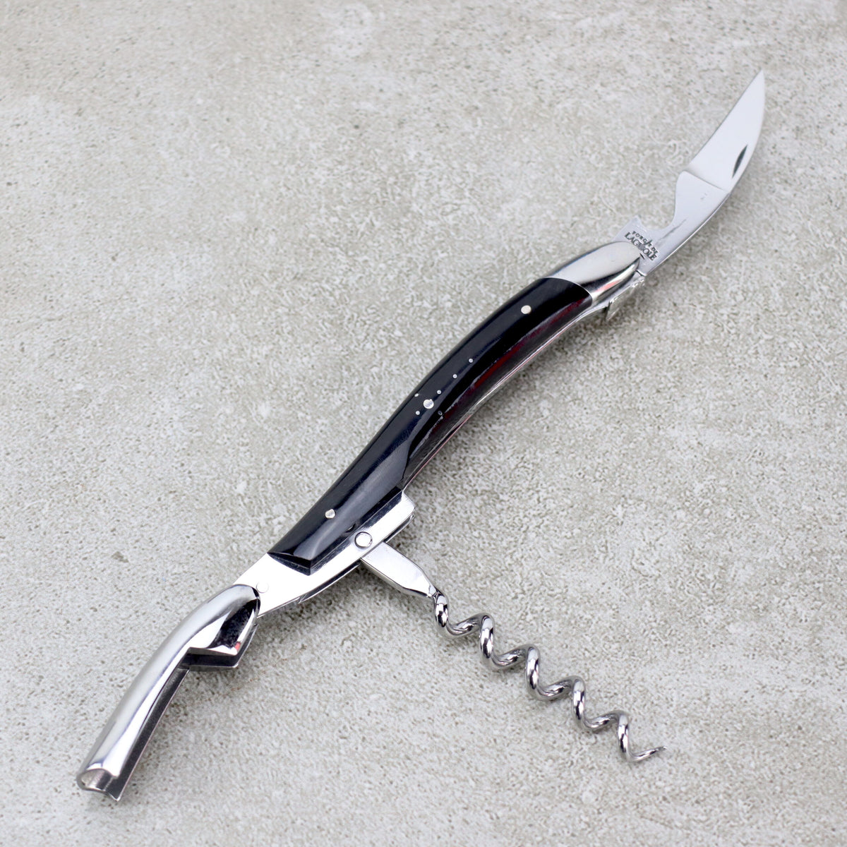Das Forge De Laguiole Kellnermesser Sommelier | Hornspitze glänzend ist ein multifunktionales Werkzeug mit einer gebogenen Messerklinge, einem ausklappbaren Korkenzieher und einem Flaschenöffner. Der schlanke Griff weist dunkle und metallische Akzente auf und verkörpert die Handwerkskunst von Forge De Laguiole. Dieses exquisite Sommelier-Werkzeug ruht auf einer grauen, strukturierten Oberfläche und ist damit eine unverzichtbare Ergänzung für die Sammlung eines jeden Sommeliers.