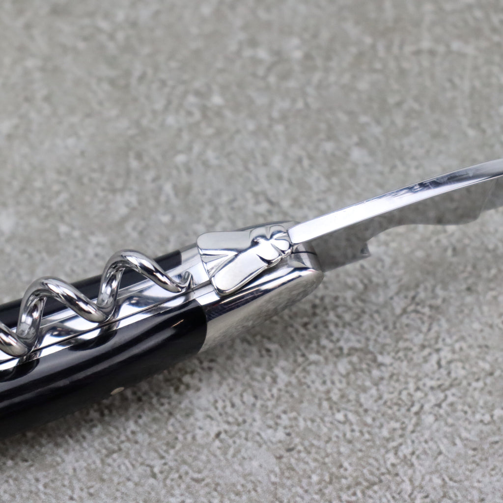 Nahaufnahme des Forge De Laguiole Kellnermessers Sommelier | Hornspitze glänzend, mit einem Korkenzieher aus Metall mit dekorativem Griff. Der spiralförmige Schraubenteil ist neben einer kleinen, dünnen Klinge zum Schneiden von Folie sichtbar. Der glänzende, polierte Griff hebt sich vom strukturierten grauen Hintergrund ab.