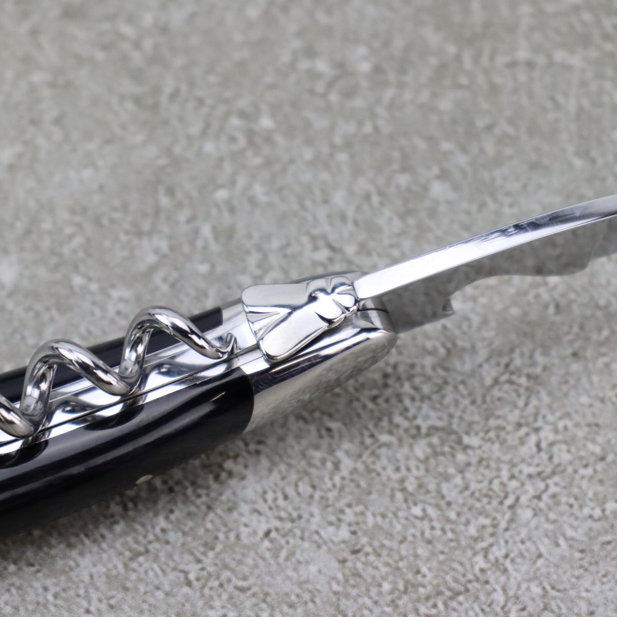 Nahaufnahme des Forge De Laguiole Kellnermessers Sommelier | Hornspitze glänzend, mit einem Korkenzieher aus Metall mit dekorativem Griff. Der spiralförmige Schraubenteil ist neben einer kleinen, dünnen Klinge zum Schneiden von Folie sichtbar. Der glänzende, polierte Griff hebt sich vom strukturierten grauen Hintergrund ab.