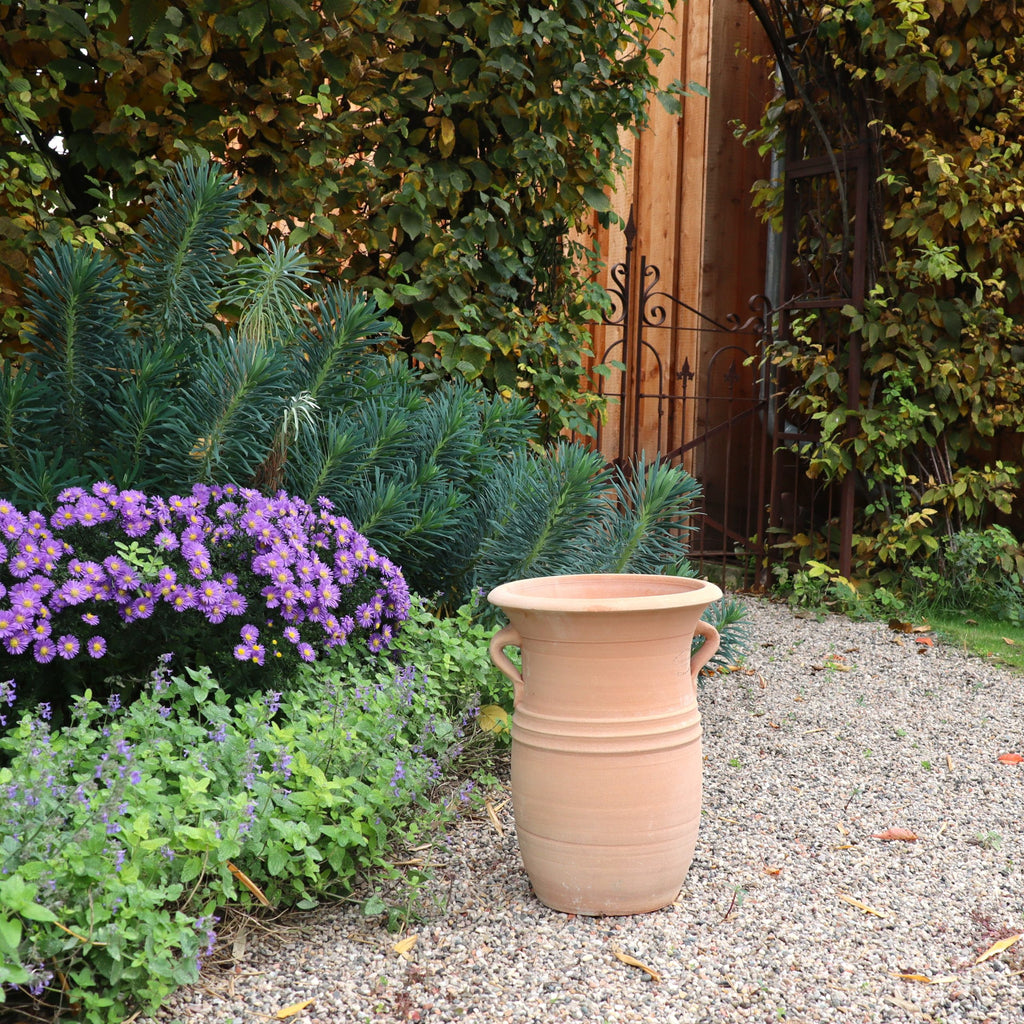 Die Vase aus Terracotta "Cistus 2", H=40cm, steht auf einem Kiesweg neben grünen Sträuchern und blühenden violetten Blumen. Im Hintergrund sind ein Holzzaun und ein Metall-Gartenbogen zu sehen.