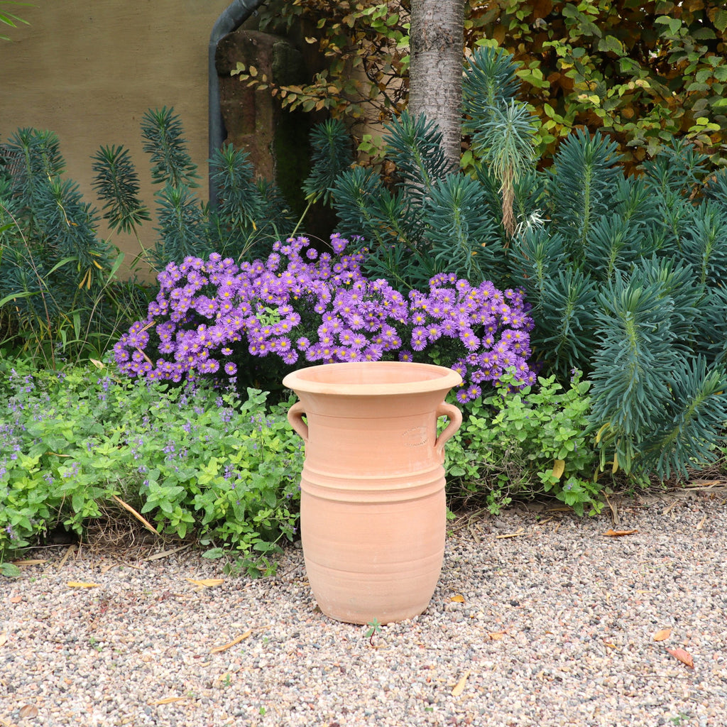 Die Vase aus Terracotta "Cistus 2" (H=40cm) mit Henkeln steht auf Kies, umgeben von üppig grünem Laub und blühenden lila Blumen in einer Gartenlandschaft.