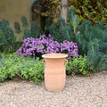 Die Vase aus Terracotta "Cistus 2" (H=40cm) mit Henkeln steht auf Kies, umgeben von üppig grünem Laub und blühenden lila Blumen in einer Gartenlandschaft.