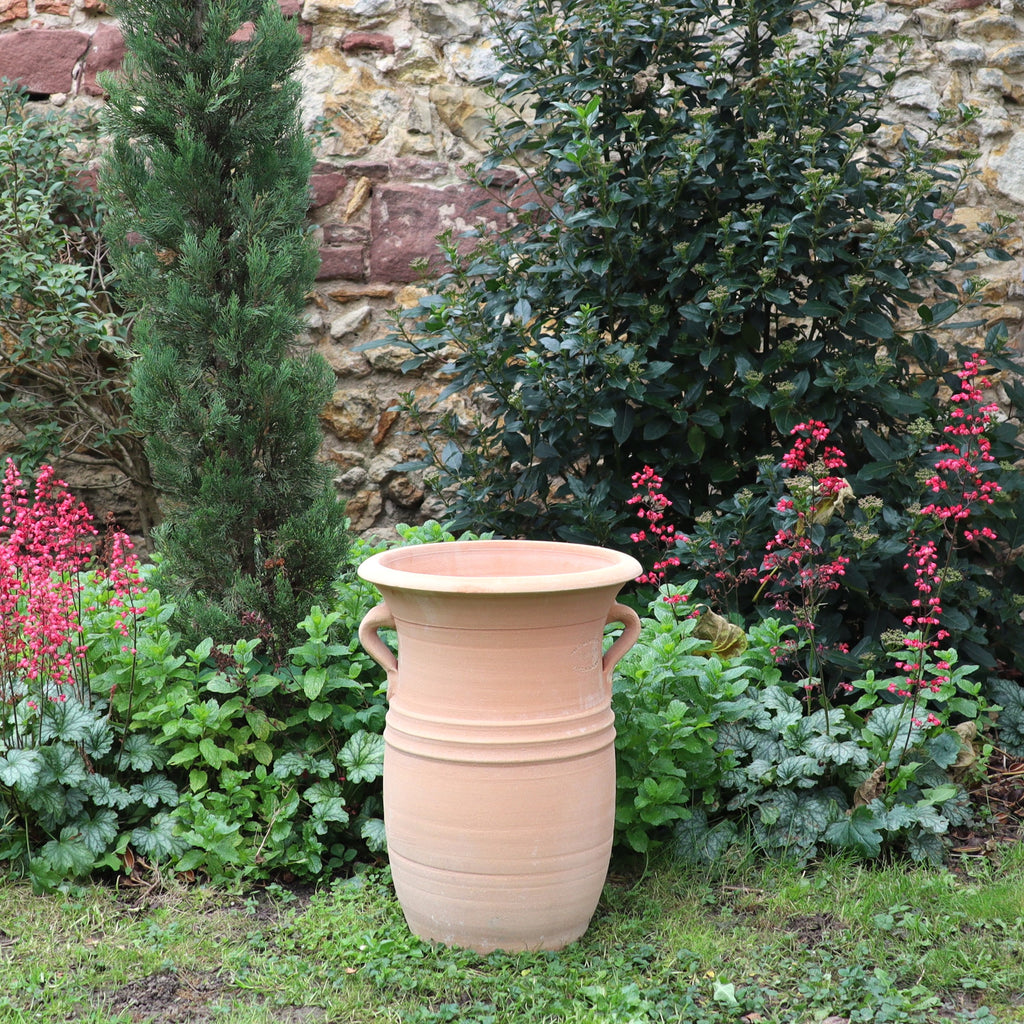 Die Vase aus Terracotta "Cistus 2" (H=40cm) mit Henkeln steht im Garten, umgeben von grünem Laub, rot blühenden Pflanzen, einem kleinen Baum und einer Steinmauer im Hintergrund.