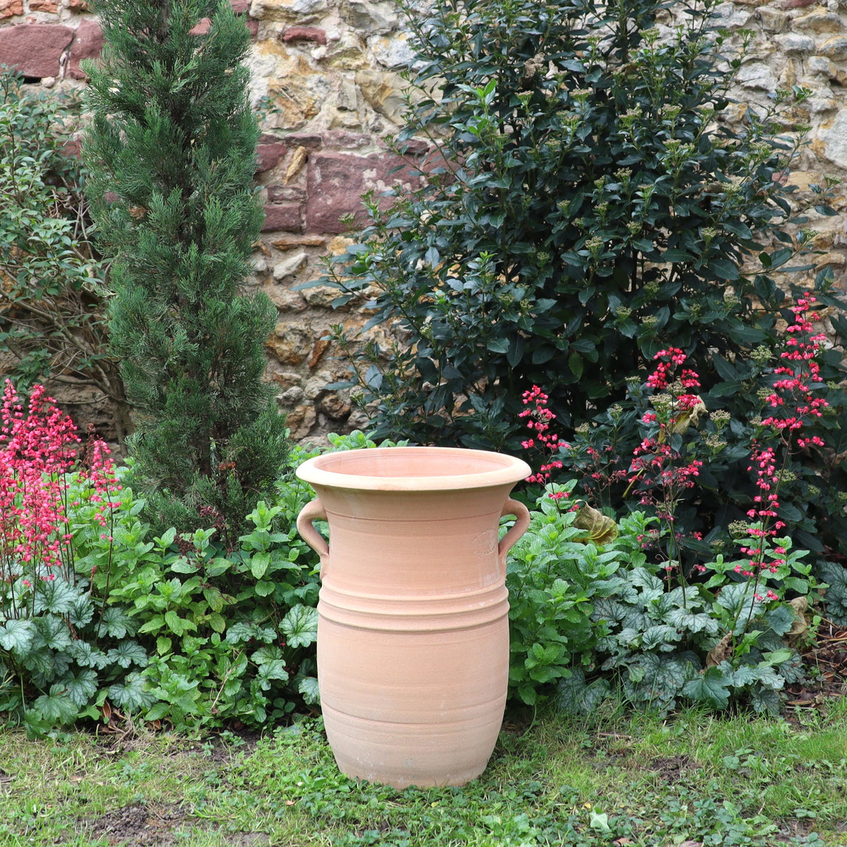 Die Vase aus Terracotta "Cistus 2" (H=40cm) mit Henkeln steht im Garten, umgeben von grünem Laub, rot blühenden Pflanzen, einem kleinen Baum und einer Steinmauer im Hintergrund.
