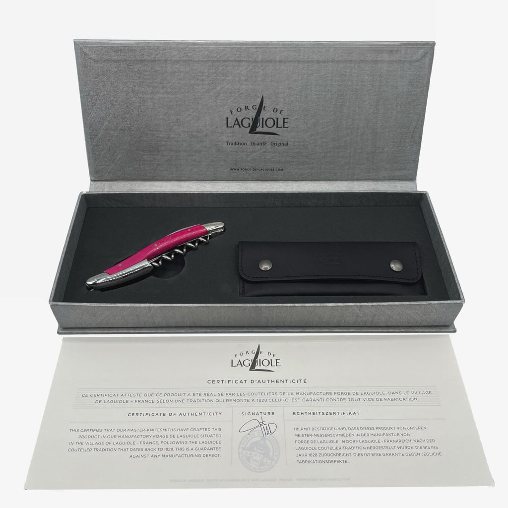 Ein hochwertiges Forge De Laguiole Kellnermesser Sommelier in rosa glänzend wird in einer offenen grauen Schachtel präsentiert, darunter befindet sich ein schwarzes Lederetui. Im unteren Teil des Bildes ist ein Echtheitszertifikat abgebildet, im Deckel der Schachtel sind das Logo und die Marke „Forge de Laguiole“ sichtbar.