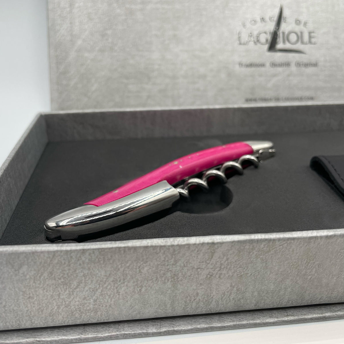 Das Forge De Laguiole Kellnermesser Sommelier in Rosa glänzend mit rosa Griff und metallisch-silberner Spitze mit Spirale liegt in einer eleganten offenen grauen Box mit der Prägung „Forge de Laguiole“ auf der Innenseite des Deckels. Die gut beleuchtete Umgebung bringt die polierte Oberfläche des Korkenziehers wunderschön zur Geltung.