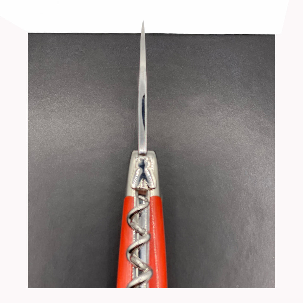 Nahaufnahme, Draufsicht eines offenen Korkenziehers und Messers des Forge De Laguiole Kellnermesser Sommelier | Orange glänzend, erinnert an die Eleganz der Designs von Forge De Laguiole. Die Messerklinge zeigt nach oben, während der Korkenzieher ausgefahren ist. Der Hintergrund ist in einer einfarbigen dunklen Farbe gehalten.