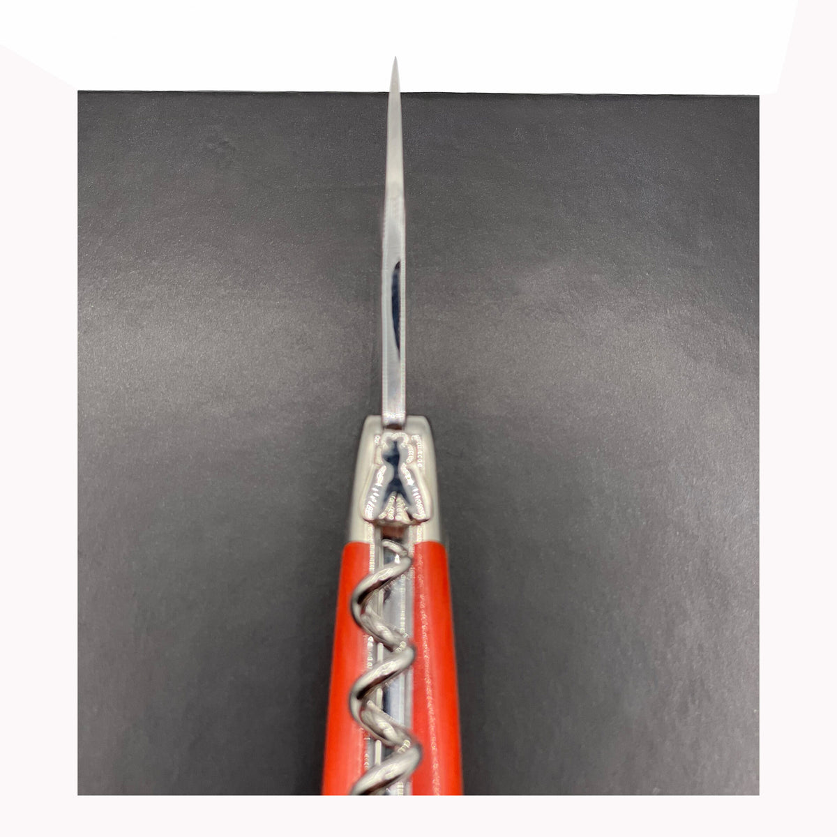 Nahaufnahme, Draufsicht eines offenen Korkenziehers und Messers des Forge De Laguiole Kellnermesser Sommelier | Orange glänzend, erinnert an die Eleganz der Designs von Forge De Laguiole. Die Messerklinge zeigt nach oben, während der Korkenzieher ausgefahren ist. Der Hintergrund ist in einer einfarbigen dunklen Farbe gehalten.