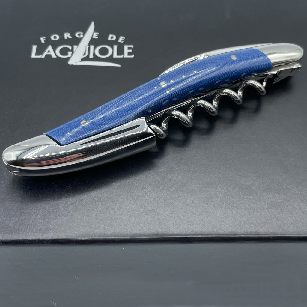 Auf einer schwarzen Oberfläche ist ein silber-blauer Korkenzieher mit polierter Oberfläche zu sehen. Der Griff mit der Aufschrift „Forge De Laguiole Kellnermesser Sommelier | Blau glänzend“ mit kleinen dekorativen Punkten ist mit dem Markennamen „Forge de Laguiole“ in Weiß über dem Korkenzieher aufgedruckt, was ihn zu einer eleganten Ergänzung für das Werkzeugset eines jeden Sommeliers macht.