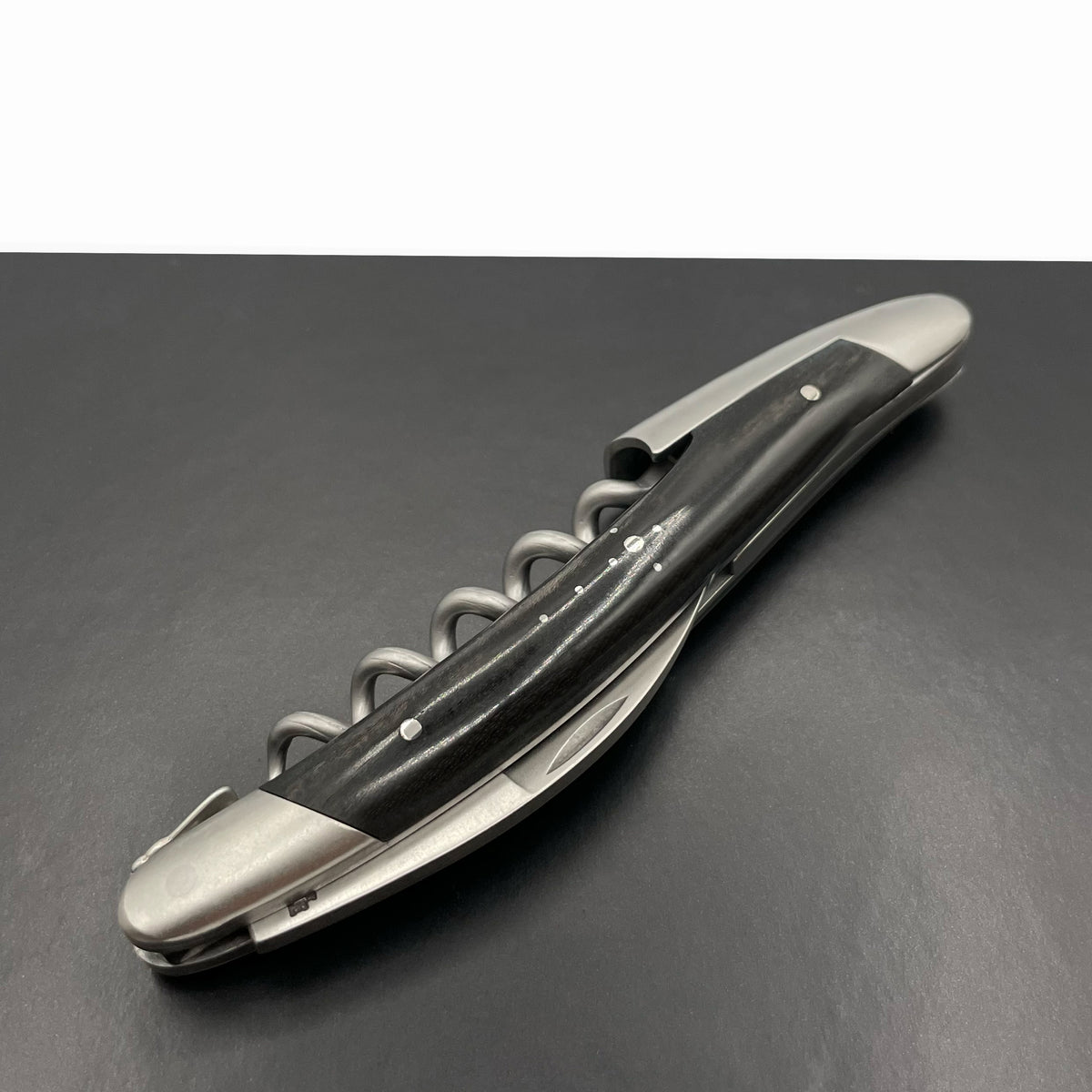 Ein geschlossenes Forge De Laguiole Kellnermesser Sommelier | Ebenholz matt, mit mattem schwarz-silbernem Design, liegt flach auf einer dunklen Oberfläche. Die Metallspirale und zusätzliche Werkzeuge sind sauber in den schlanken schwarzen Griff eingearbeitet und bieten Präzision auf Sommelier-Niveau.