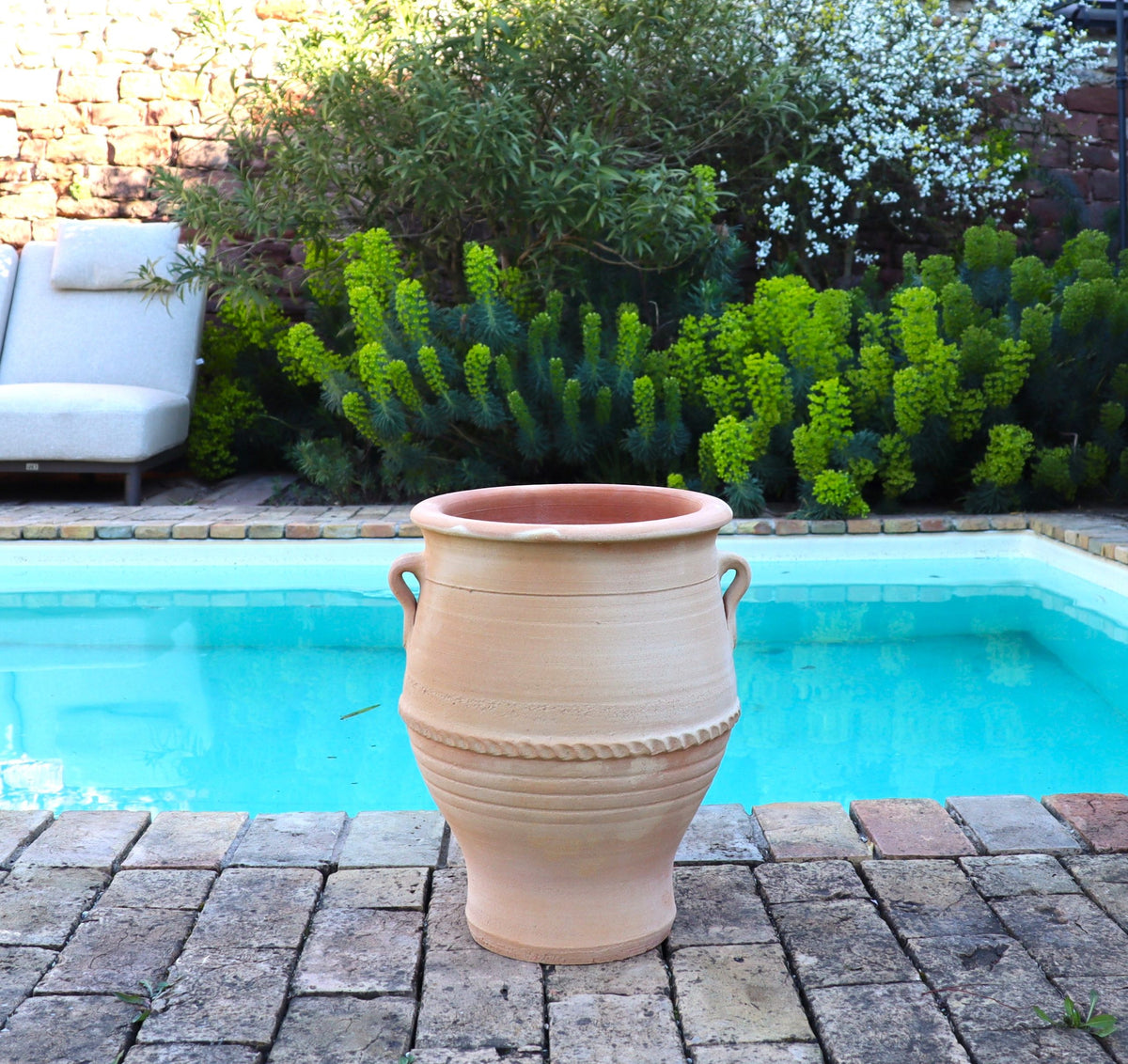 Eine frostfeste Terrakotta Amphore "Thymus 2" H=40cm mit zwei Griffen steht auf einer gemauerten Terrasse am Pool, umgeben von grünen Sträuchern, blühenden Pflanzen und einem Liegestuhl im Hintergrund.