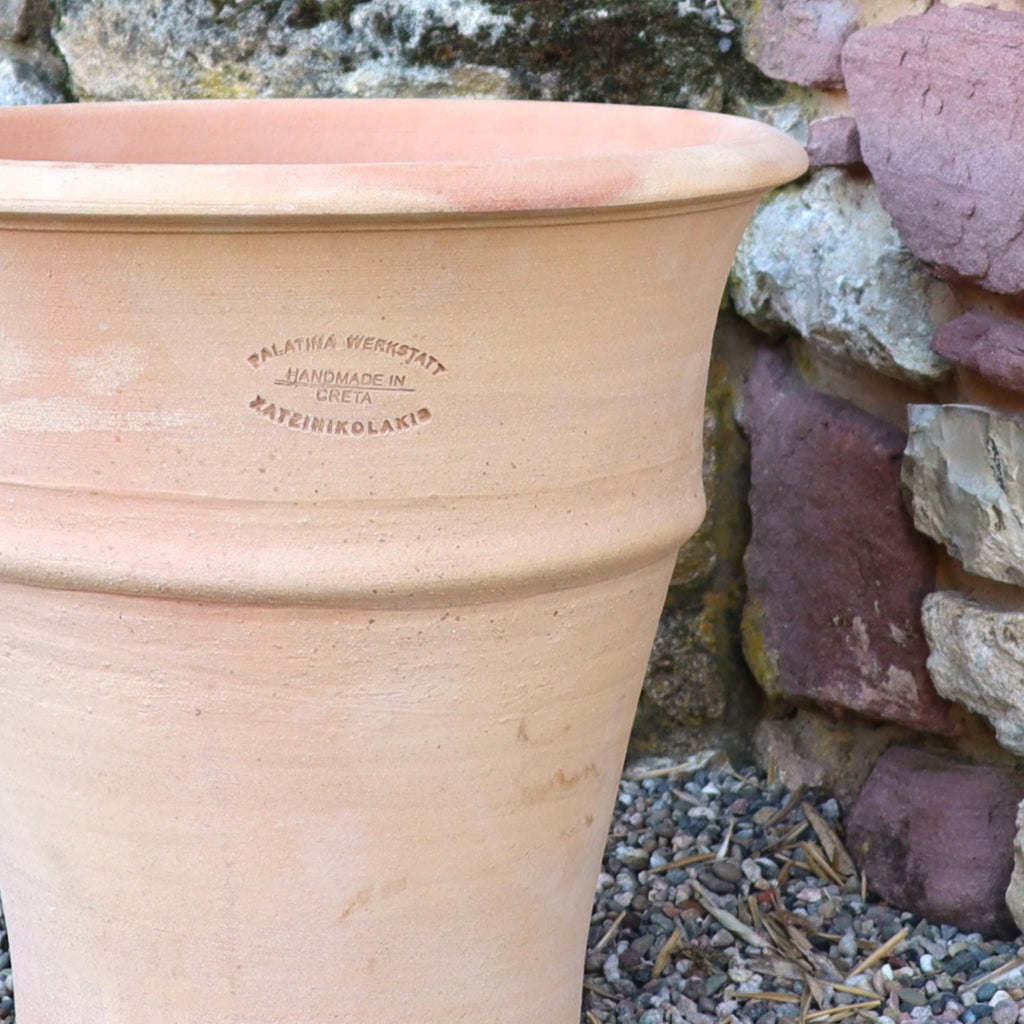 Das winterharte Terrakotta-Pflanzgefäß "Yucca", H=35 cm, mit geprägtem Etikett "PALATINA WERKSTATT HANDMADE IN CRETA KATSEFIKOLAKIS", steht auf Kies neben einer Steinmauer.