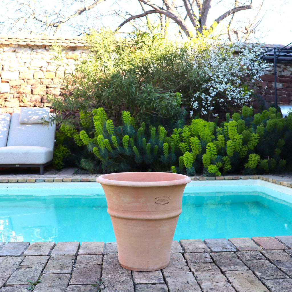 Das winterharte Terrakotta-Pflanzgefäß "Yucca", H=35 cm, steht auf Ziegelsteinpflaster an einem Pool, umgeben von grünen Sträuchern und einer Steinmauer. Auf der linken Seite sind zwei Liegestühle teilweise sichtbar. Blühende Sträucher bringen Farbe in den Hintergrund.