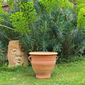 Das winterharte Terracotta Pflanzgefäß "Rosa", H=35 cm, steht auf dem Rasen vor üppigem Grün, links daneben eine Steinskulptur mit teilweise herausgearbeitetem menschlichen Gesicht.