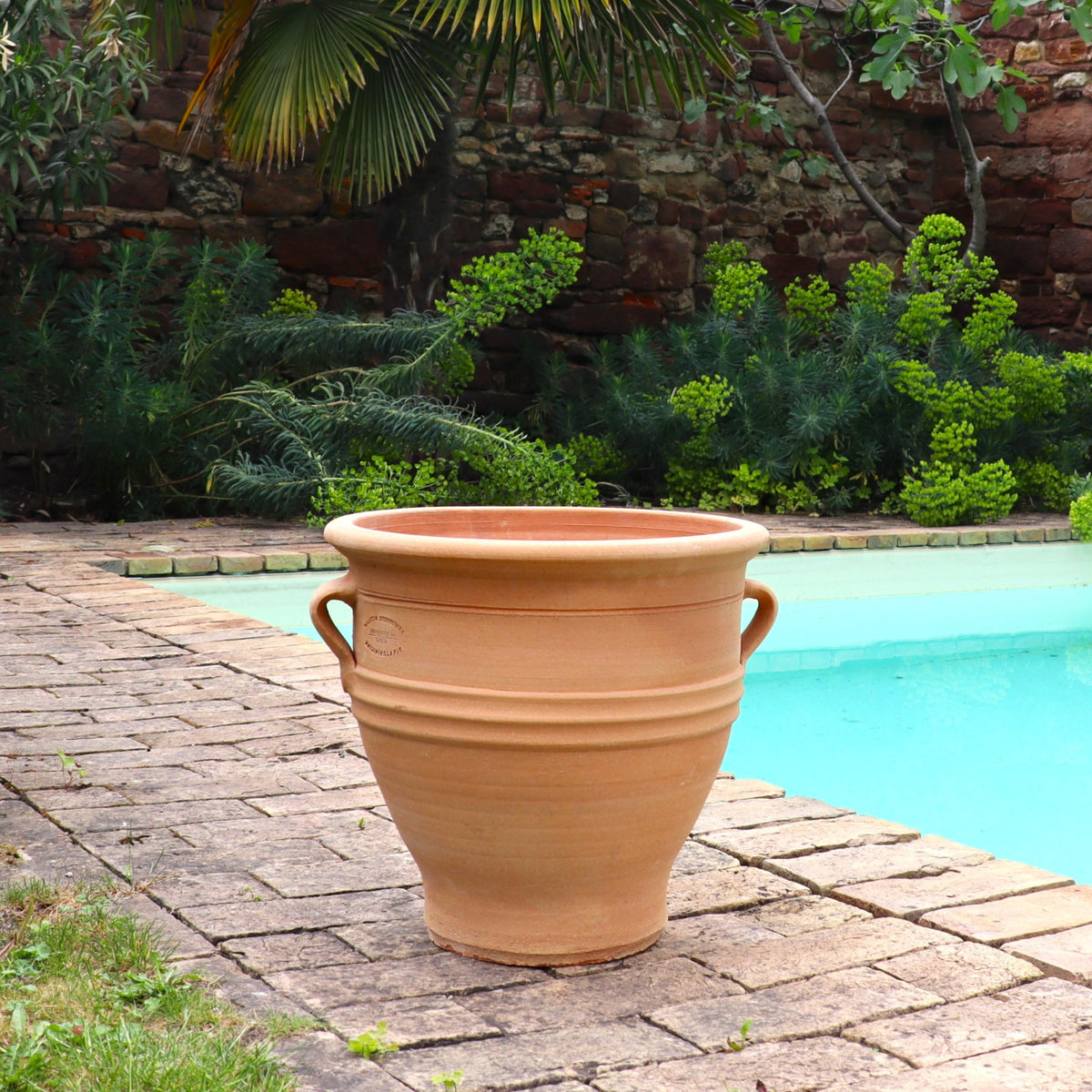 Ein winterhartes Terracotta-Pflanzgefäß "Rosa", H=35 cm, mit Henkeln steht auf einer gemauerten Terrasse neben einem türkisfarbenen Pool, umgeben von üppigen Grünpflanzen und einer Steinmauer im Hintergrund.
