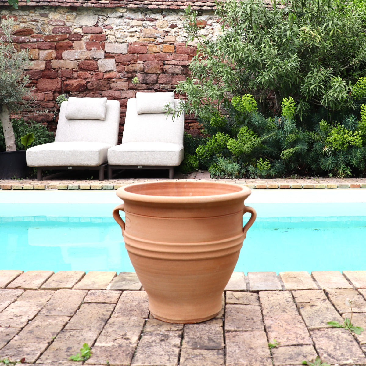 Das winterharte Terracotta Pflanzgefäß "Rosa", H=35 cm, steht auf Ziegelpflaster am türkisfarbenen Pool, flankiert von zwei gepolsterten Liegestühlen und üppigem Grün vor einer rustikalen Steinmauer.