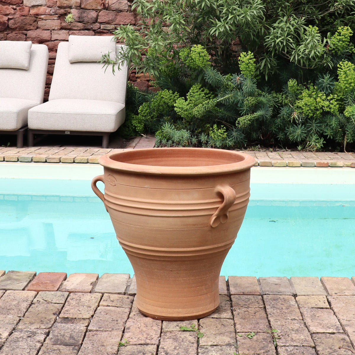 Das Terrakotta-Pflanzgefäß "Rosa", H=40 cm (Kopie) mit Henkeln steht auf einer gemauerten Terrasse neben einem Pool, umgeben von grünen Sträuchern und zwei gepolsterten Liegestühlen, die einen ruhigen Rückzugsort im Garten schaffen.