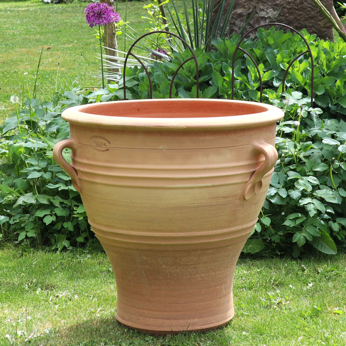 Das Terrakotta-Pflanzgefäß "Rosa", H=40 cm (Kopie), hat zwei seitliche Griffe und horizontale Rillen. Es ist auf Gras in einem Garten mit üppigem Grün und lila Blumen im Hintergrund dargestellt.