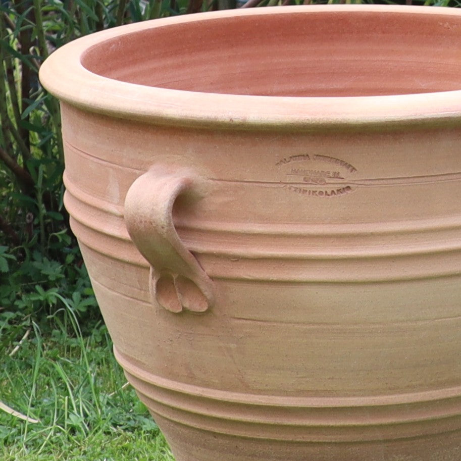 Das Terrakotta-Pflanzgefäß "Rosa", H=40 cm (Kopie), mit dekorativem Henkel und horizontalen Rillen, steht auf dem Rasen inmitten von üppigem Gartenlaub; in der Nähe des Randes befindet sich eine gestempelte Herstellermarke.