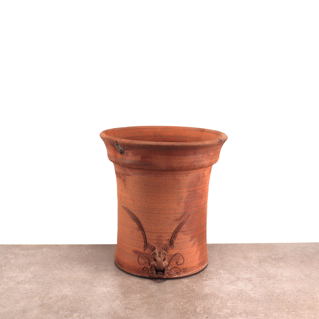 Ein rustikaler, zylindrischer Blumentopf aus Terrakotta mit leicht ausgestelltem Rand und seitlichen Griffen. Der frostsichere Blumentopf hat eine glatte Oberfläche und verfügt über ein dekoratives Design am Boden, das an den Kopf eines gehörnten Tieres erinnert und auf einer flachen, neutral gefärbten Oberfläche ruht.