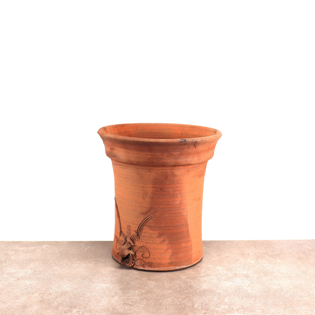Ein Blumentopf Blumenkübel aus Terrakotta, ein frostbeständiger Blumentopf, steht auf einer Betonoberfläche. Der Topf weist ein einfaches Blumenmuster mit geprägten Blättern und Wirbeln auf der Vorderseite auf. Der Hintergrund ist schlicht weiß und lenkt die Aufmerksamkeit voll auf das rustikale Aussehen des Topfes.